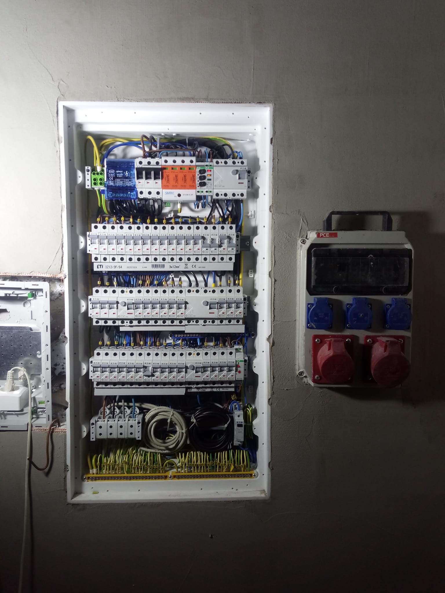 Rozdzielnica elektryczna z zabezpieczeniami nadprądowymi i różnicowoprądowymi, bezpiecznikami, przewodami w kolorach oraz gniazdami siłowymi na ścianie z surowego betonu.
