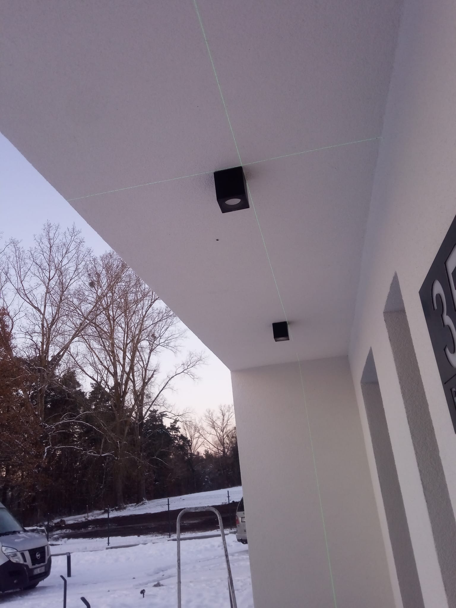 Dwa czarne, kwadratowe reflektory zewnętrzne zamontowane na białym podbitce w Goleniowie, wyrównane laserowo, widok z dołu na tle zimowego krajobrazu.