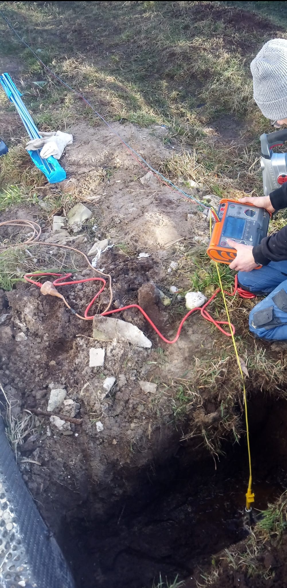 Pomiar rezystancji uziomu w wykopie, z użyciem miernika i sondy, na trawiastym terenie.