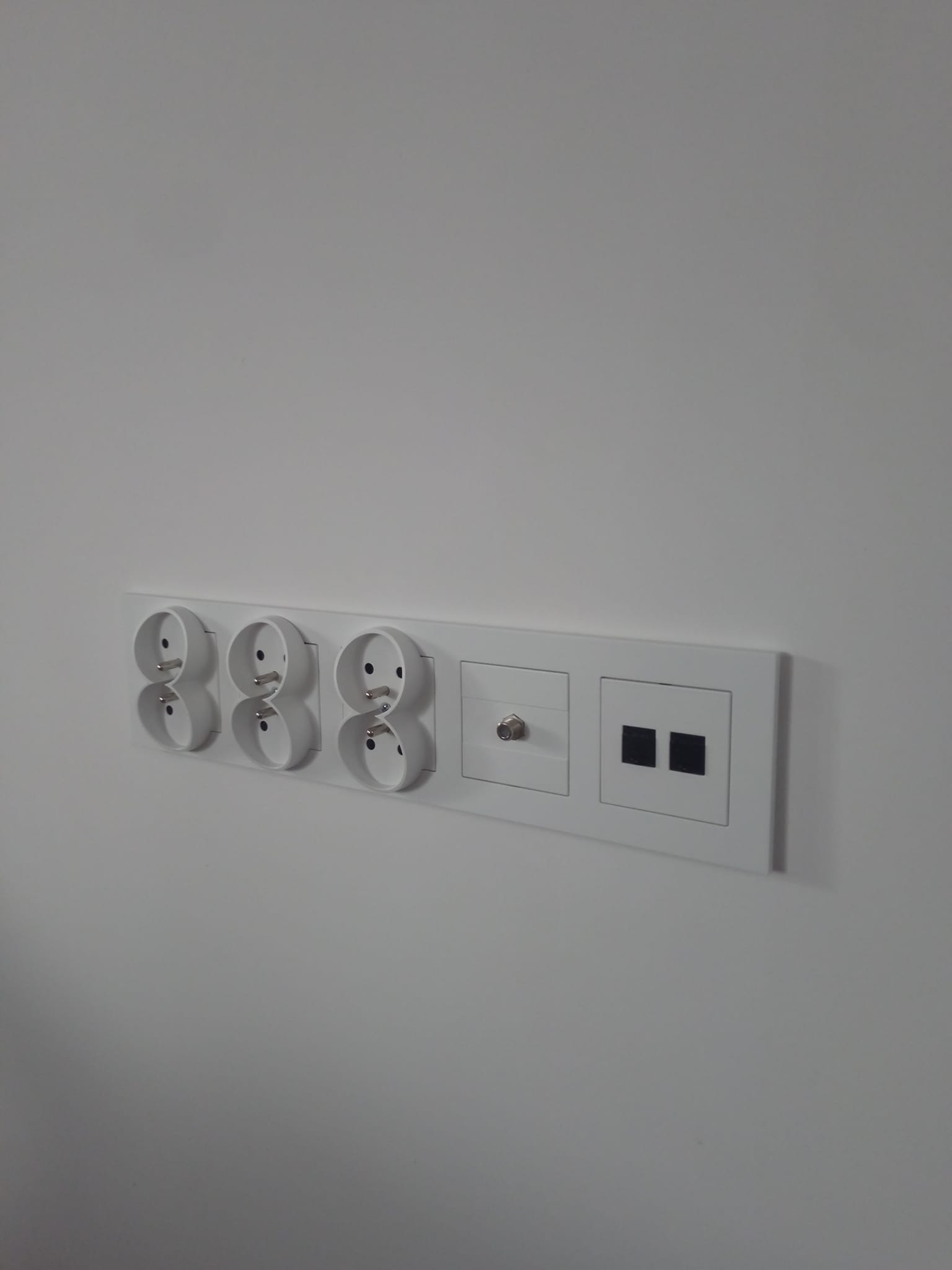 Potrójne gniazdko elektryczne, gniazdo antenowe i podwójny włącznik światła w białej ramce, zamontowane na jasnej ścianie.