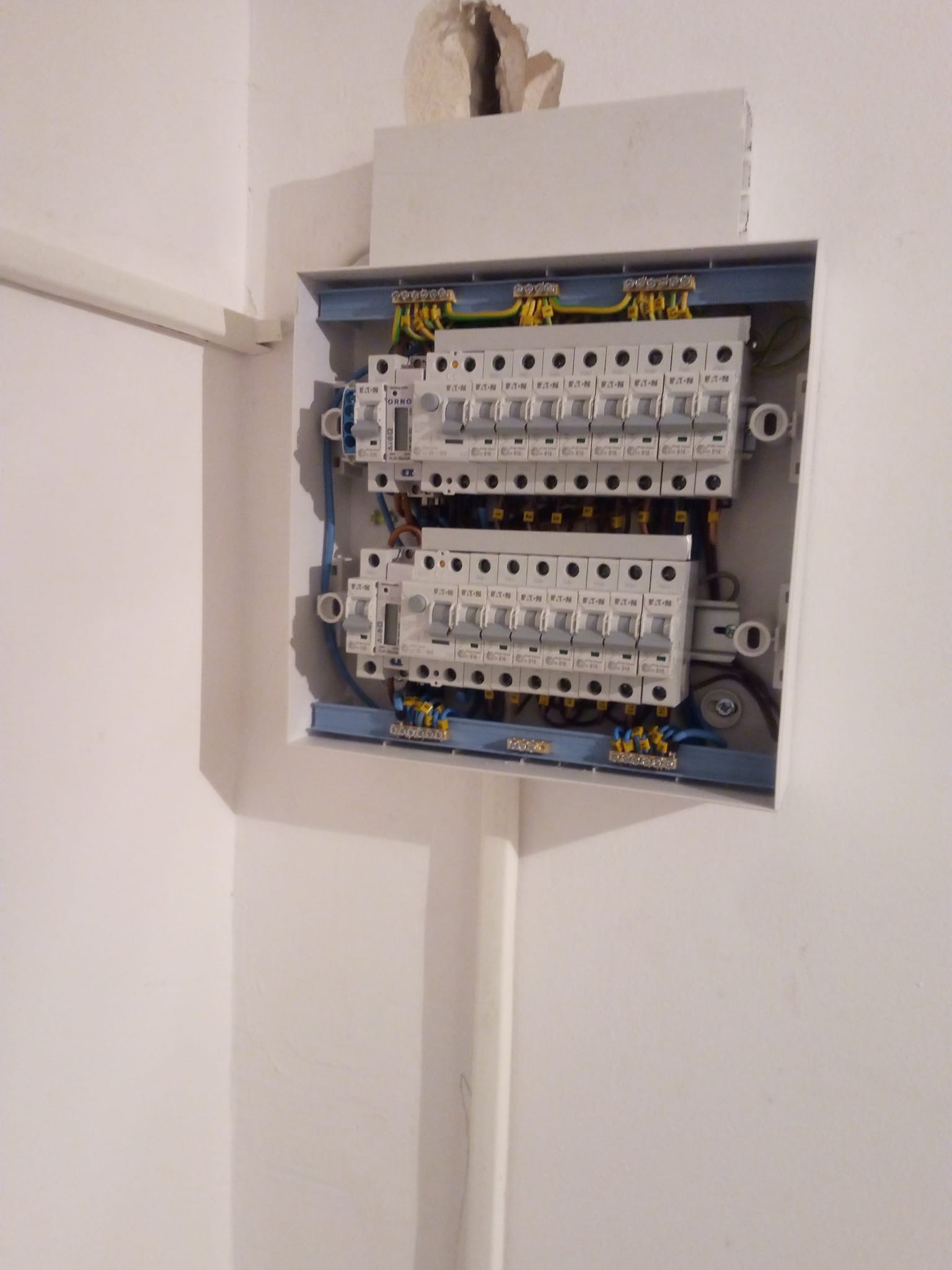 Otwarta, biała skrzynka elektryczna z dwoma rzędami bezpieczników na ścianie w trakcie montażu, widoczne kable i listwy zaciskowe, fragment ściany z ubytkiem nad skrzynką.