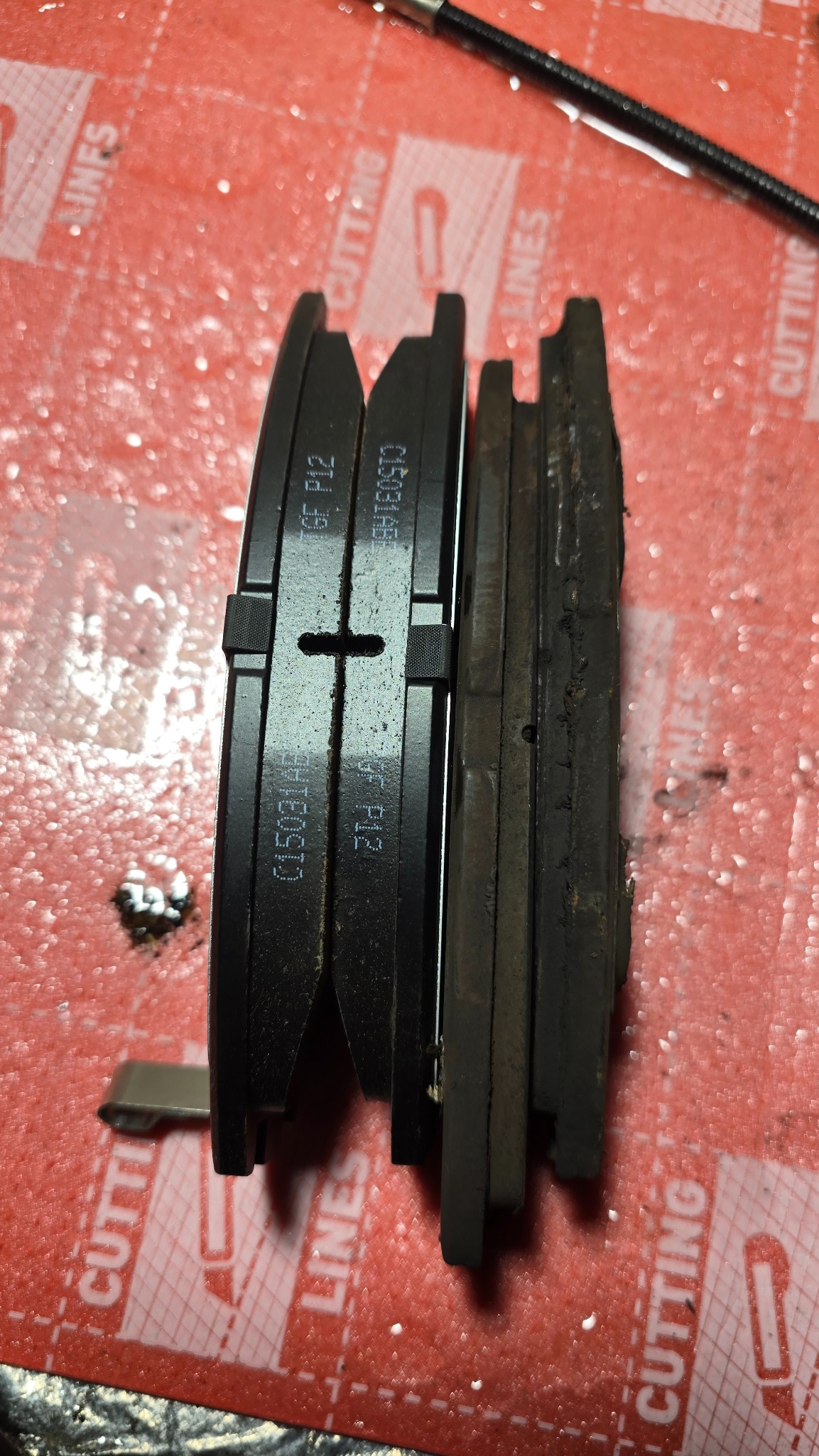 Zużyte i nowe klocki hamulcowe zestawione obok siebie na czerwonej macie. Widoczne różnice w grubości i stopniu zużycia okładzin ciernych.