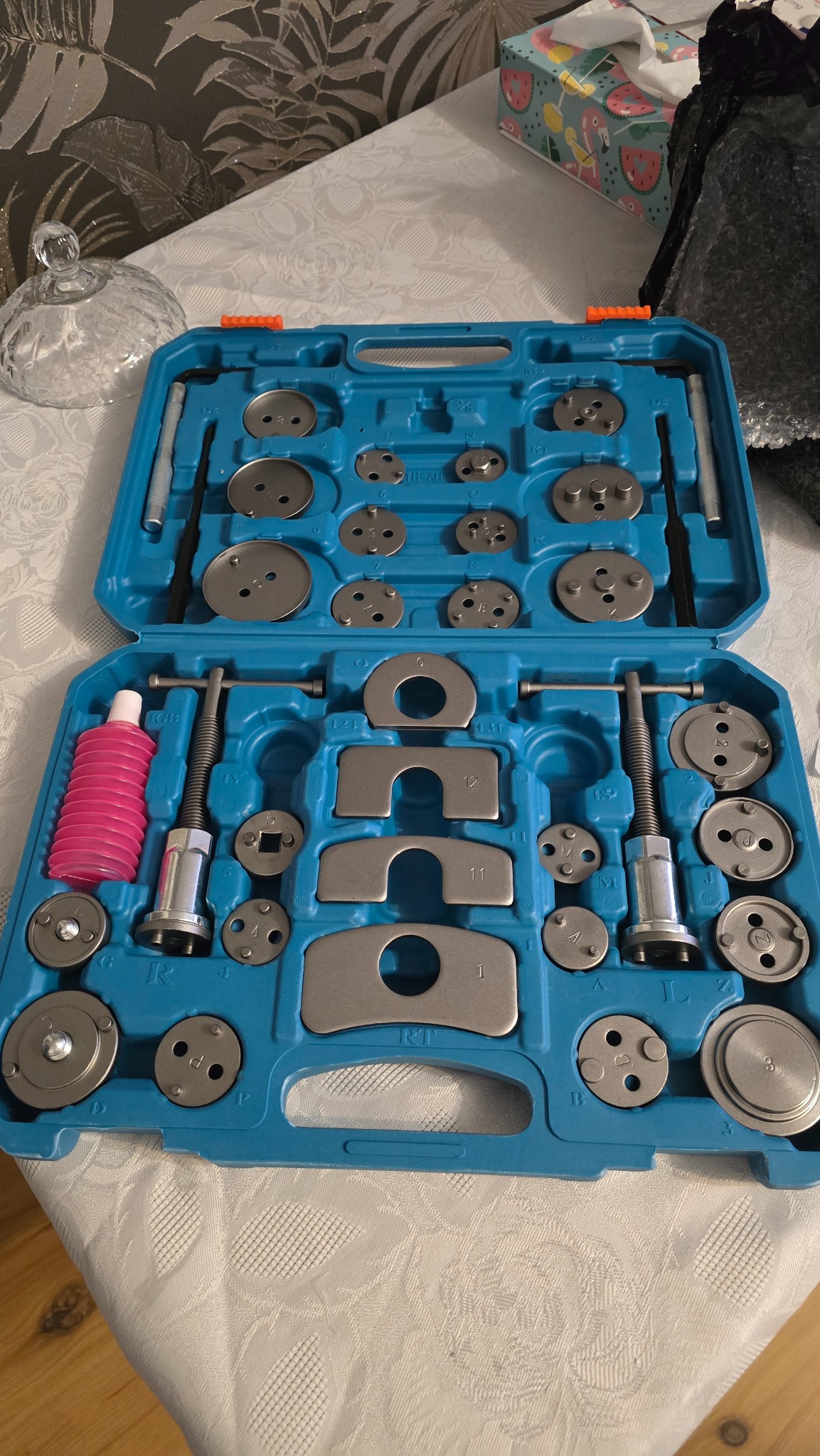 Otwarty, turkusowy zestaw narzędzi do naprawy hamulców samochodowych z różnymi adapterami i tłoczkami, leżący na białym obrusie w kwiatowy wzór.