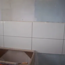 Świeżo ułożone, błyszczące, białe płytki ścienne z ciemnymi fugami w trakcie remontu, widoczna puszka elektryczna i fragment niedokończonej zabudowy szafki.