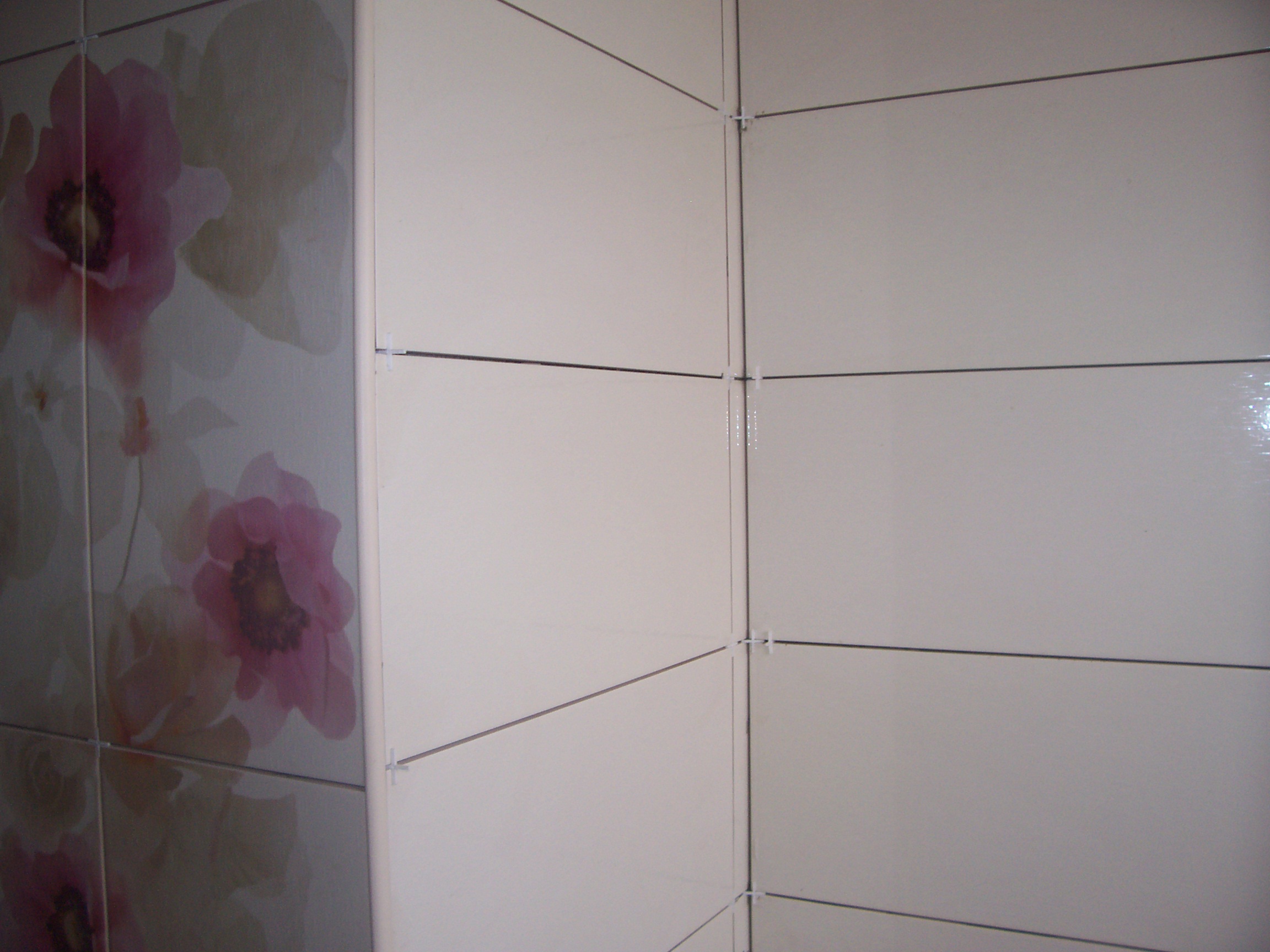 Ściana w trakcie układania płytek ceramicznych z motywem kwiatowym i białych, widoczne krzyżyki dystansowe, świeżo położone fugi.