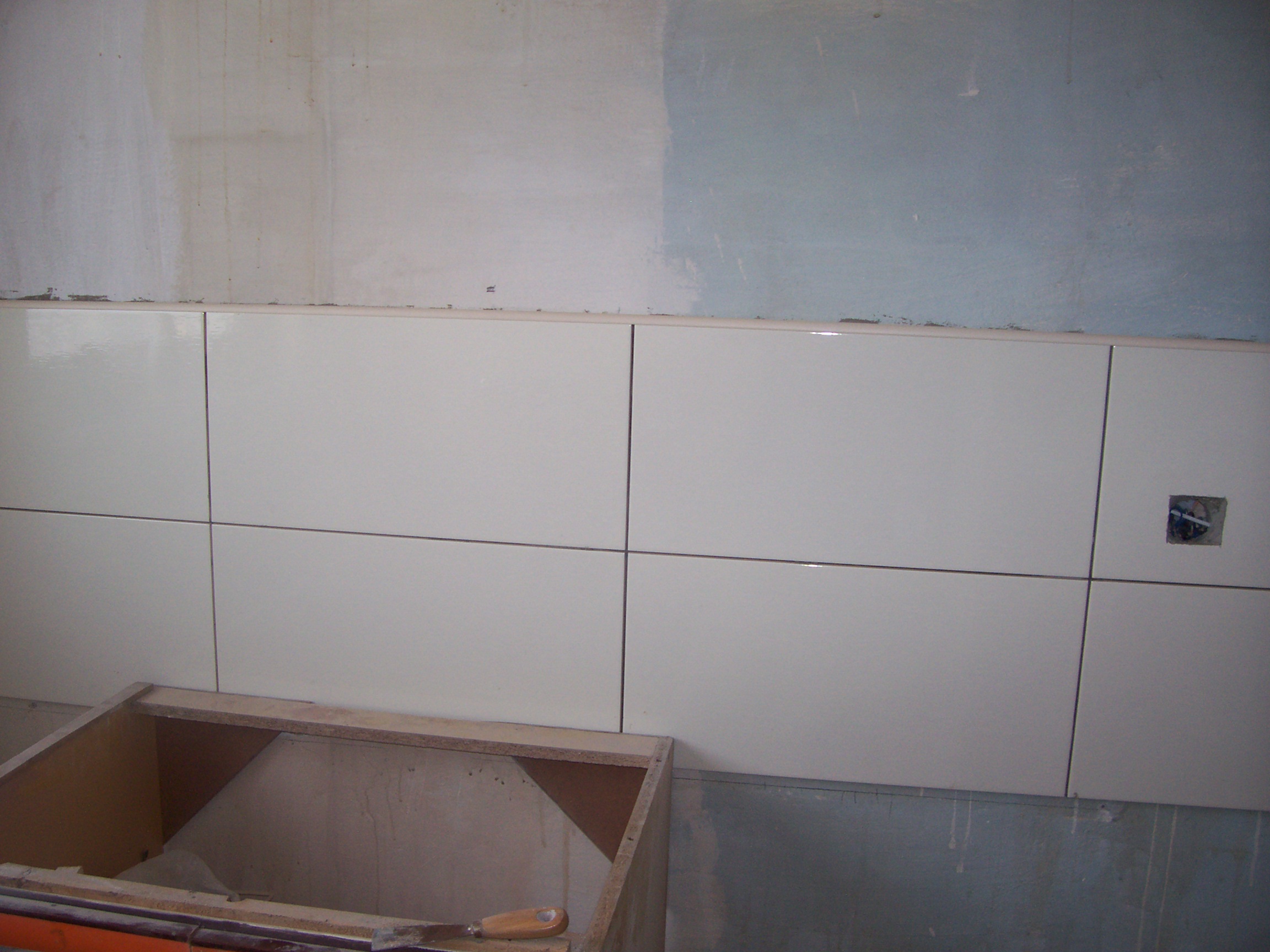 Świeżo ułożone, błyszczące, białe płytki ścienne z ciemnymi fugami w trakcie remontu, widoczna puszka elektryczna i fragment niedokończonej zabudowy szafki.