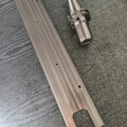 Metalowa listwa z otworami i frezowanym wycięciem oraz uchwyt narzędziowy na szarym tle, widoczne ślady obróbki.