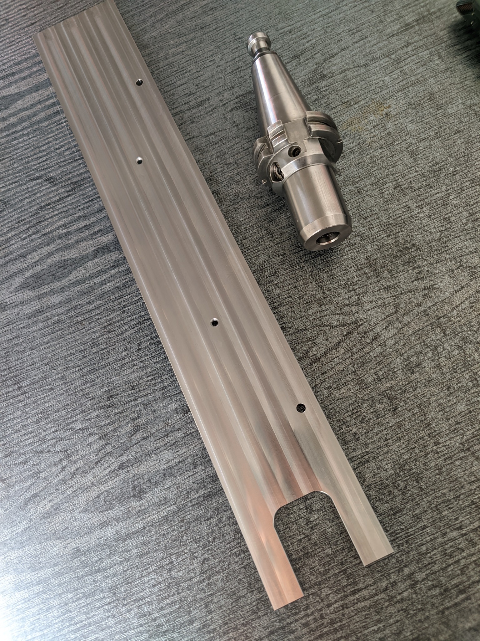 Metalowa listwa z otworami i frezowanym wycięciem oraz uchwyt narzędziowy na szarym tle, widoczne ślady obróbki.