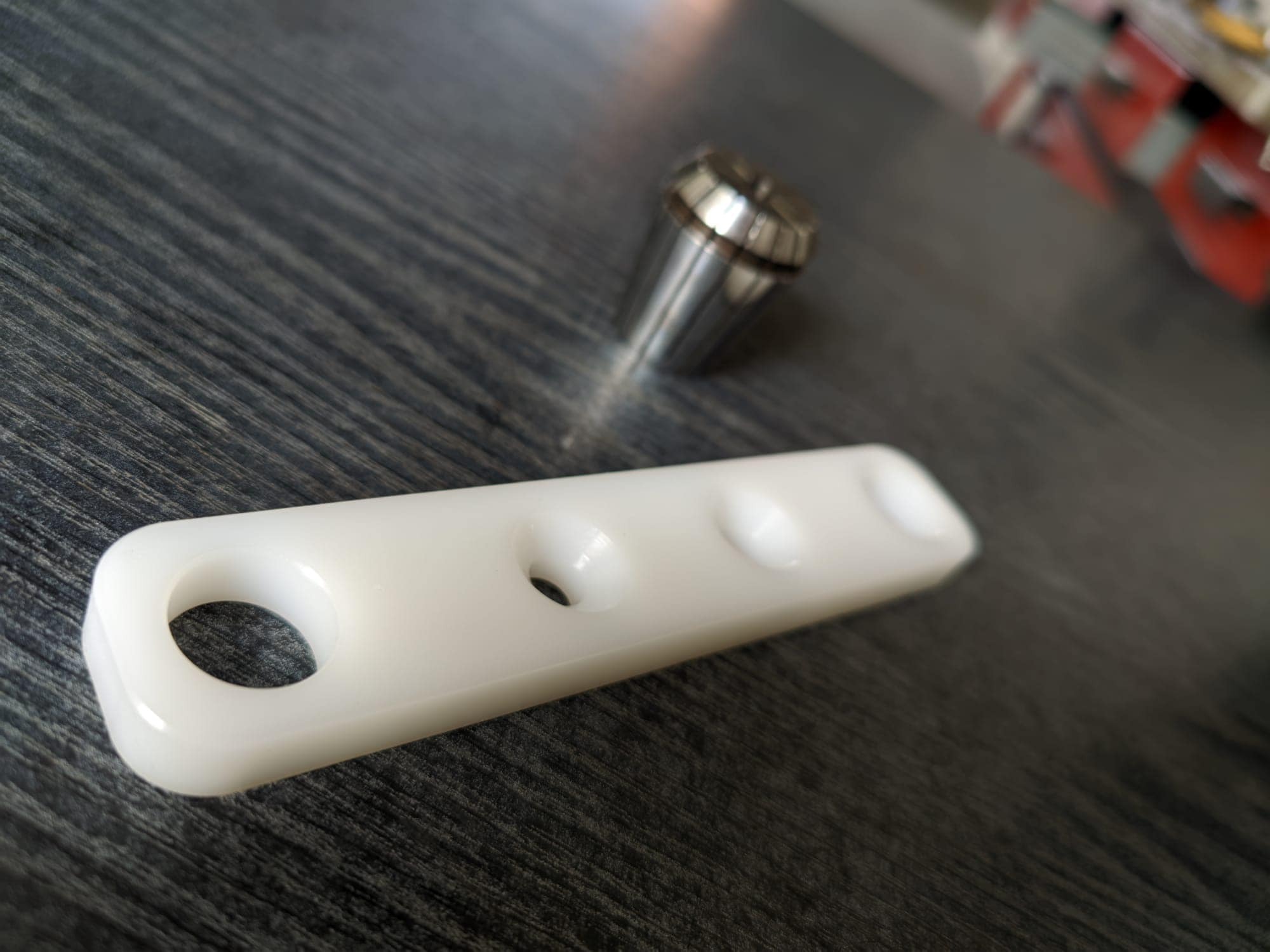 Biała, prostokątna płytka z tworzywa sztucznego z trzema otworami leżąca na ciemnym, drewnianym blacie obok metalowej tulei zaciskowej.