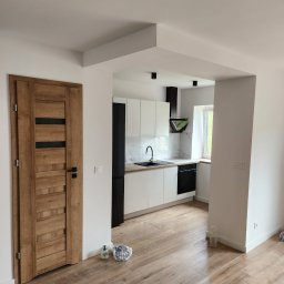 Mar-Maks - Wnętrze po remoncie: drewniane drzwi z czarnymi wstawkami, podłoga imitująca drewno, biała kuchnia z czarnymi akcentami widoczna w tle.