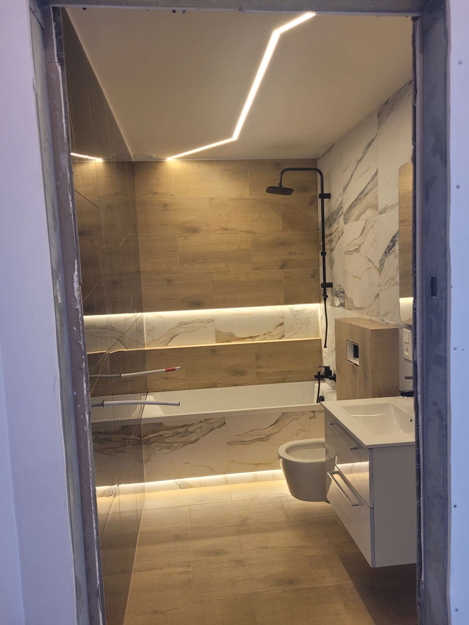 Nowoczesna łazienka z drewnopodobnymi i marmurowymi płytkami, podświetleniem LED na suficie i wzdłuż wanny, widok przez surową futrynę drzwi.