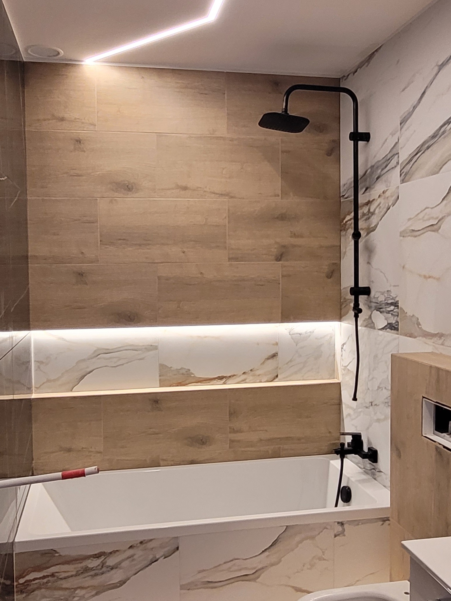 Nowoczesna łazienka w Częstochowie z wanną, czarną armaturą i podświetlanymi płytkami imitującymi drewno oraz marmur.