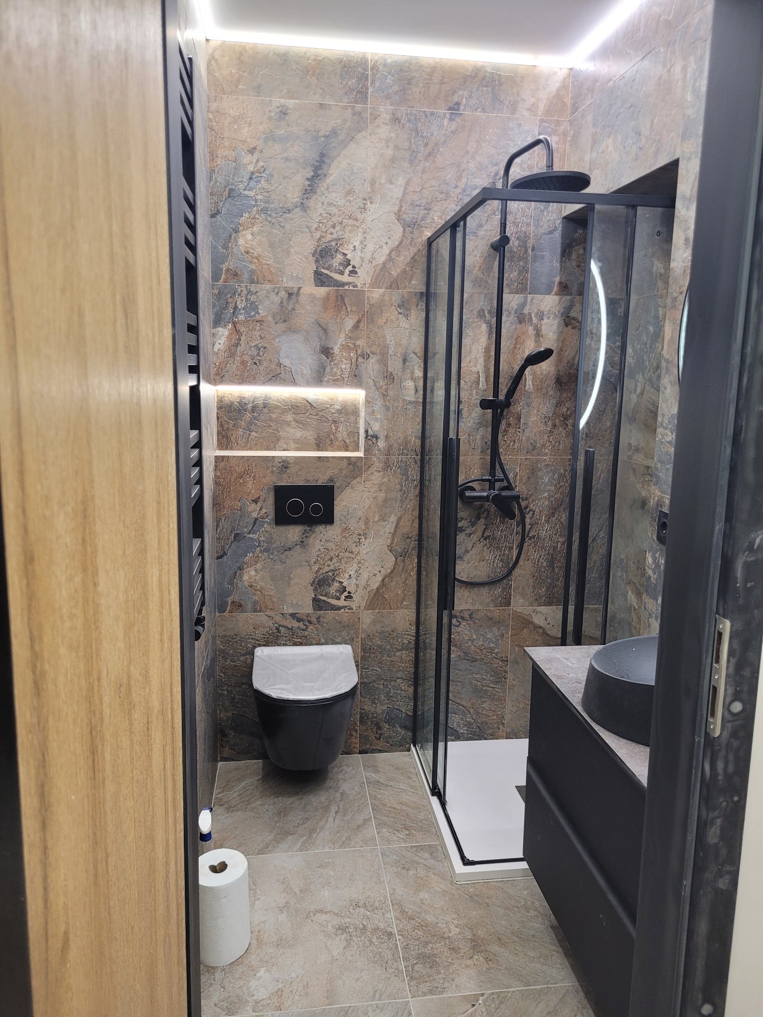 Nowoczesna łazienka z kabiną prysznicową typu walk-in z czarnymi profilami, wiszącą toaletą w kolorze czarnym z podświetlanym panelem, umywalką nablatową i płytkami imitującymi kamień.