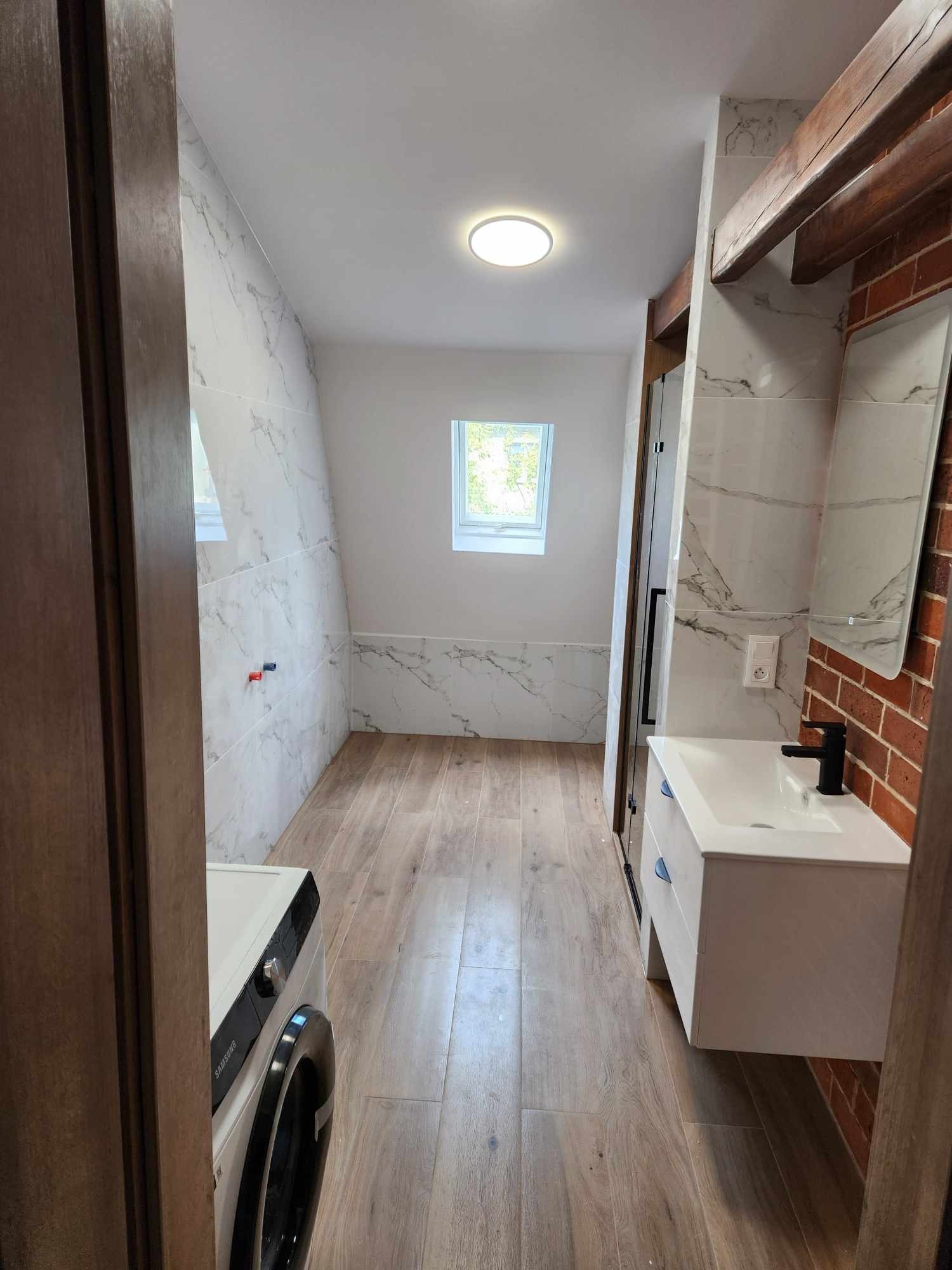 Wyremontowana łazienka na poddaszu z drewnianymi belkami, marmurowymi płytkami na ścianach, drewnianą podłogą, umywalką z czarną armaturą i pralką.