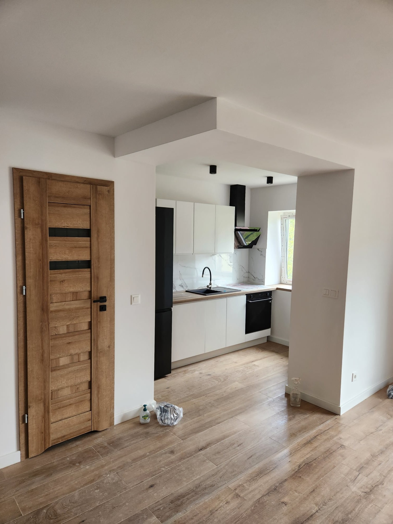 Wnętrze po remoncie: drewniane drzwi z czarnymi wstawkami, podłoga imitująca drewno, biała kuchnia z czarnymi akcentami widoczna w tle.