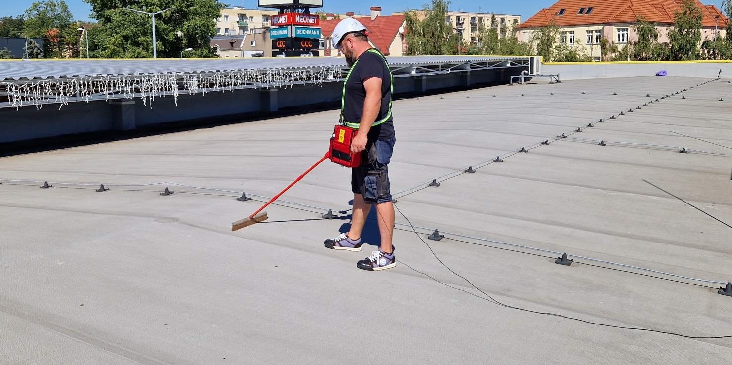 Pracownik w kasku, z uprzężą i detektorem nieszczelności dachu płaskiego w Jarużynie, sprawdza szczelność membrany dachowej za pomocą szczotki przewodzącej.