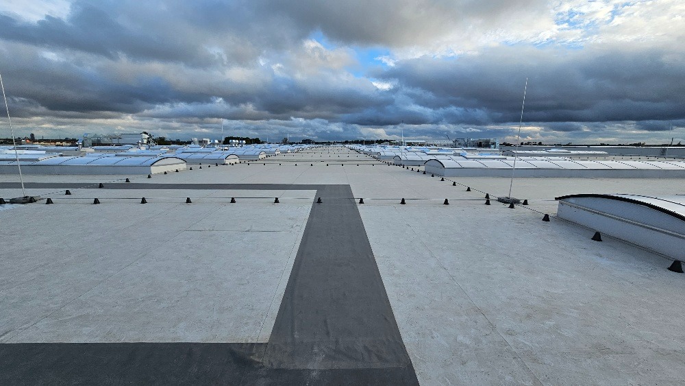 Rozległy, płaski dach przemysłowy z licznymi świetlikami i systemem odgromowym, widok z góry w pochmurny dzień.