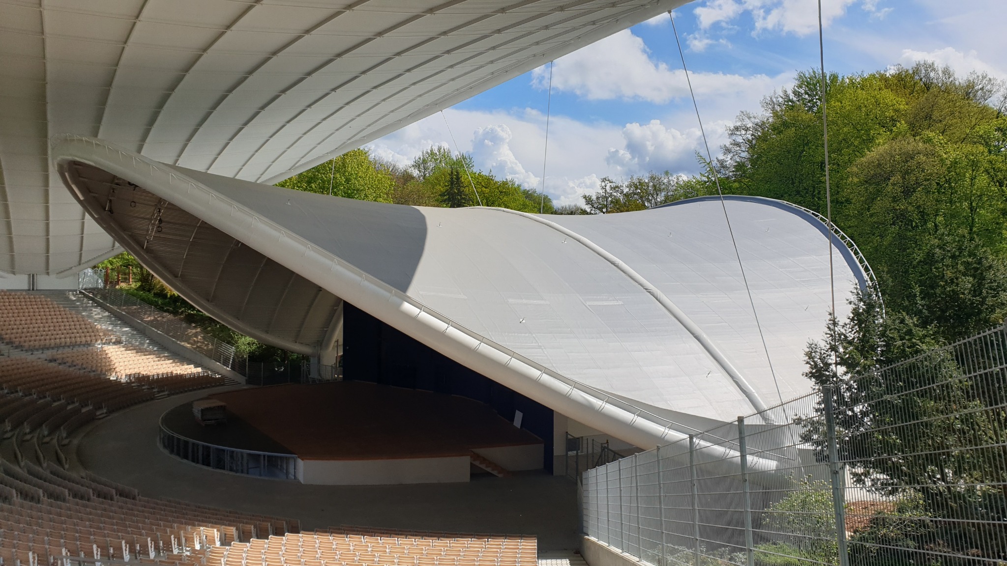 Zadaszenie amfiteatru z membraną w kształcie łuku, widok na scenę i rzędy siedzeń, tło stanowią drzewa i błękitne niebo z chmurami.
