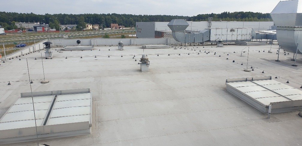 Jasnoszary dach przemysłowy z świetlikami dachowymi, wentylacją i systemem odgromowym na tle drzew i budynków.