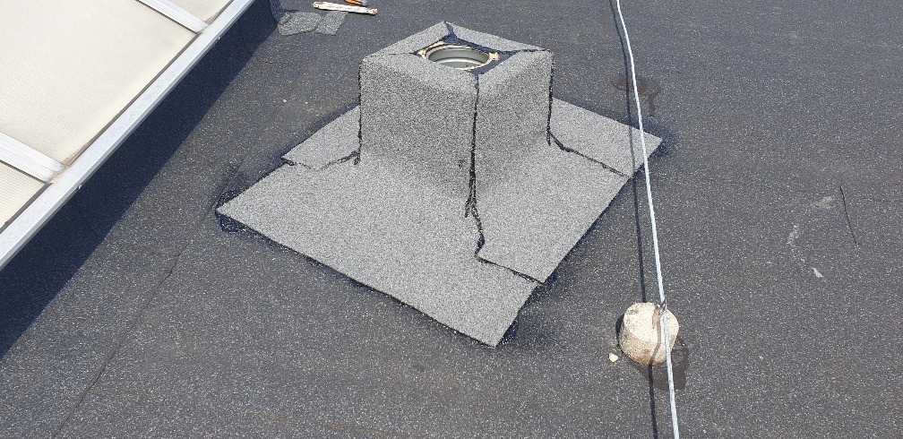 Uszczelnienie komina na płaskim dachu pokrytym papą, zabezpieczone kamieniem obciążającym linkę.