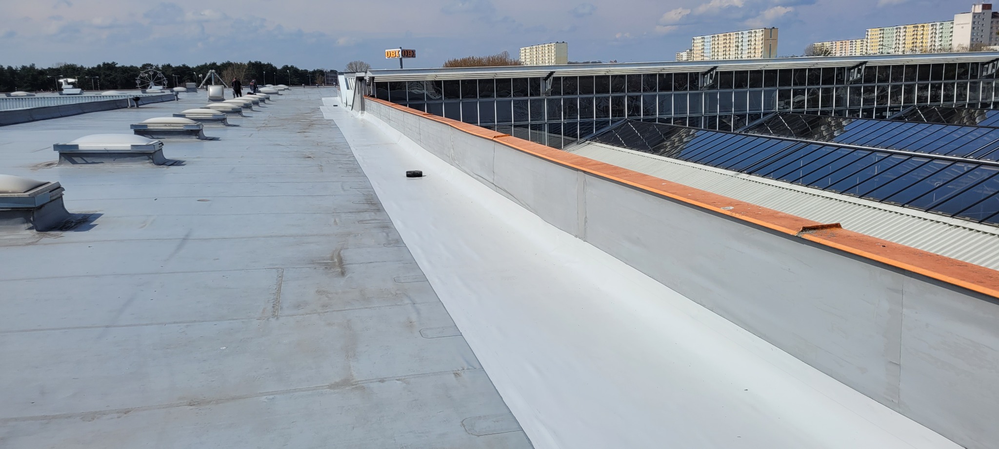 Widok na płaski dach budynku komercyjnego po zakończonej izolacji, z widocznymi świetlikami i obróbkami blacharskimi, w tle budynki mieszkalne i znak firmowy sklepu.