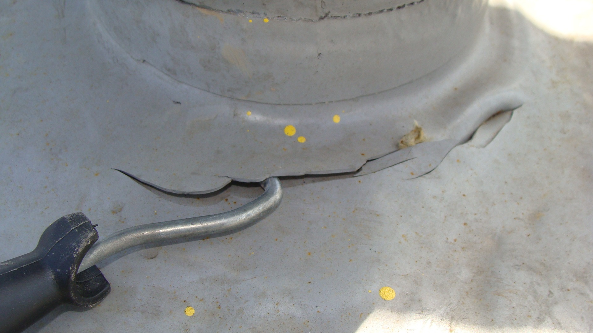 Bliskie ujęcie uszkodzonego, metalowego elementu dachu z pęknięciami i odchodzącą farbą, przy którym leży zakrzywione narzędzie z czarną rączką.