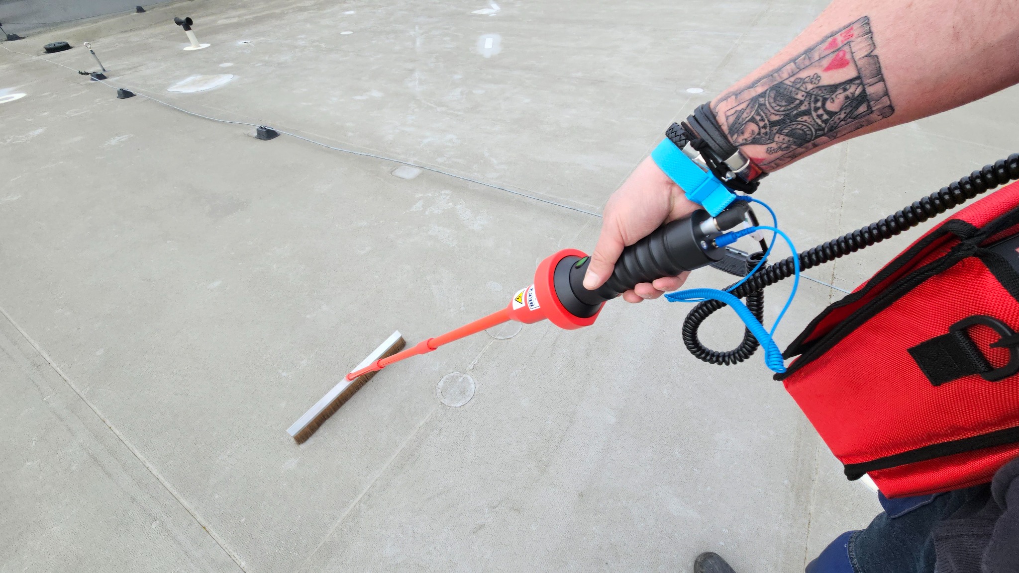 Próba szczelności dachu płaskiego z użyciem urządzenia z szczotką, widoczny tatuaż na ręce operatora.