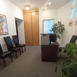 Poczekalnia w kancelarii z ciemnymi krzesłami, recepcją, roślinami doniczkowymi i plakatem 'Łódź' na ścianie.