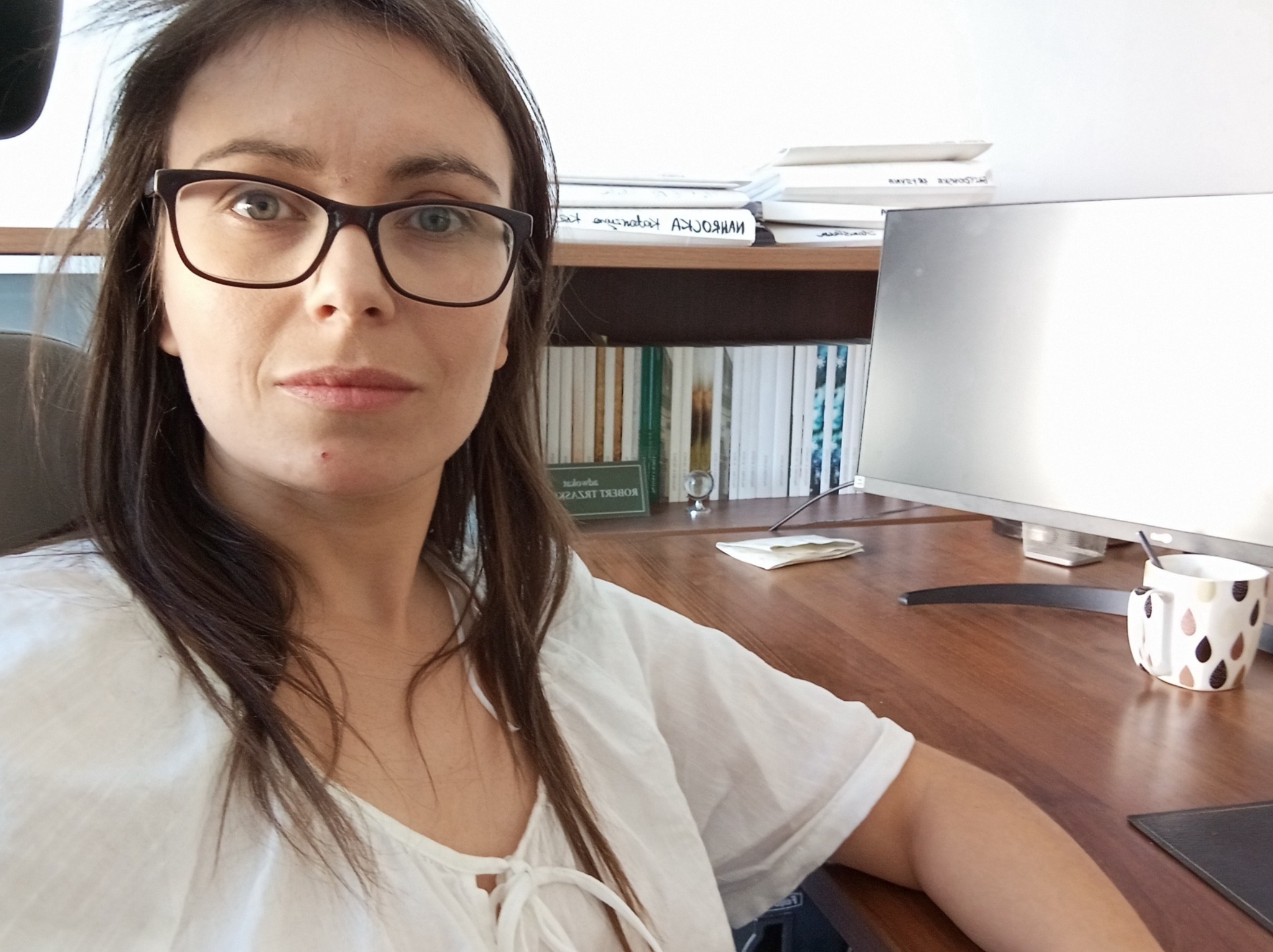 Portret kobiety w okularach siedzącej przy biurku z monitorem, książkami i kubkiem, prawdopodobnie w swoim biurze.