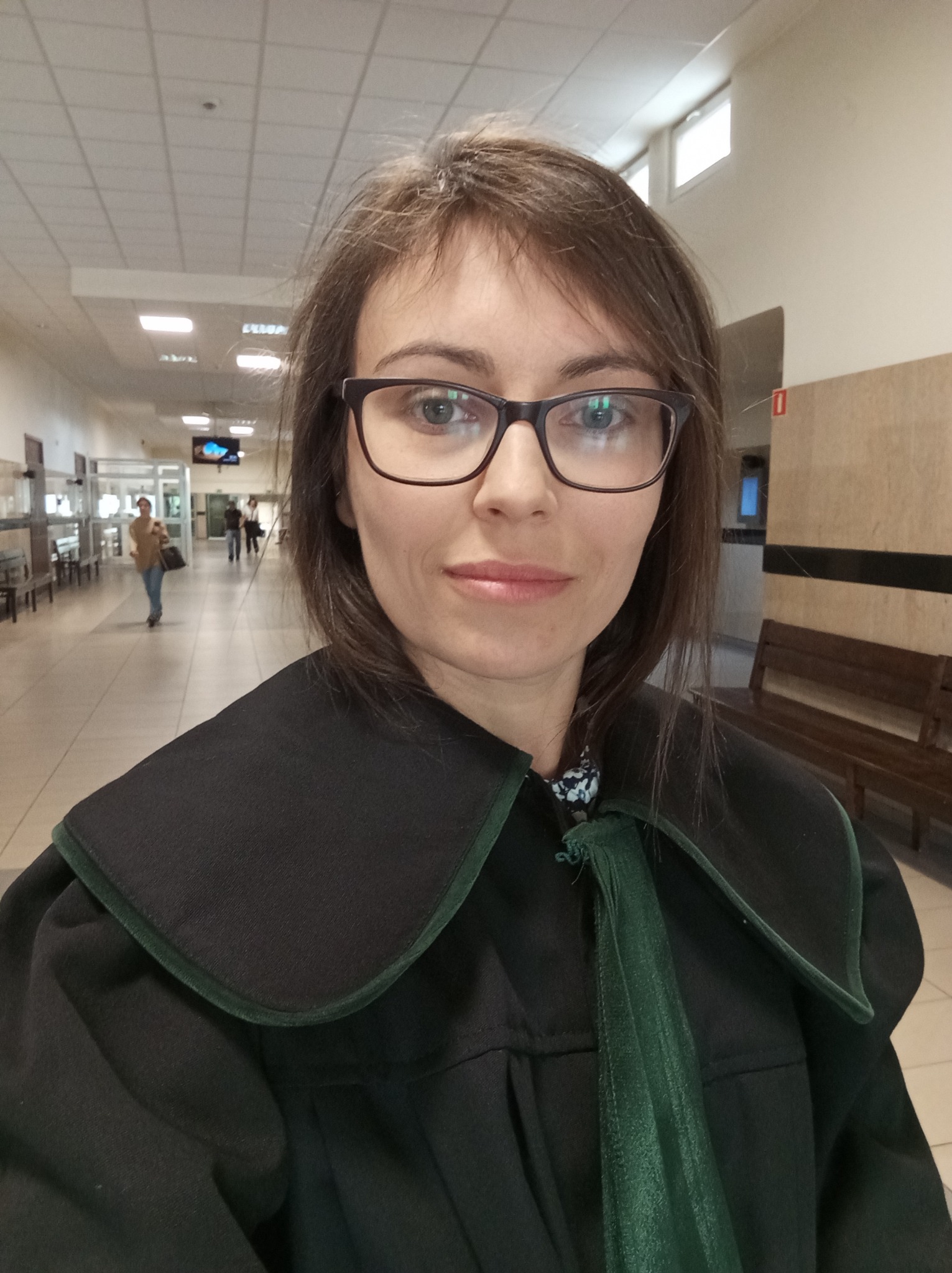 Portret kobiety w todze adwokackiej, w tle korytarz sądowy z przechodzącymi ludźmi.