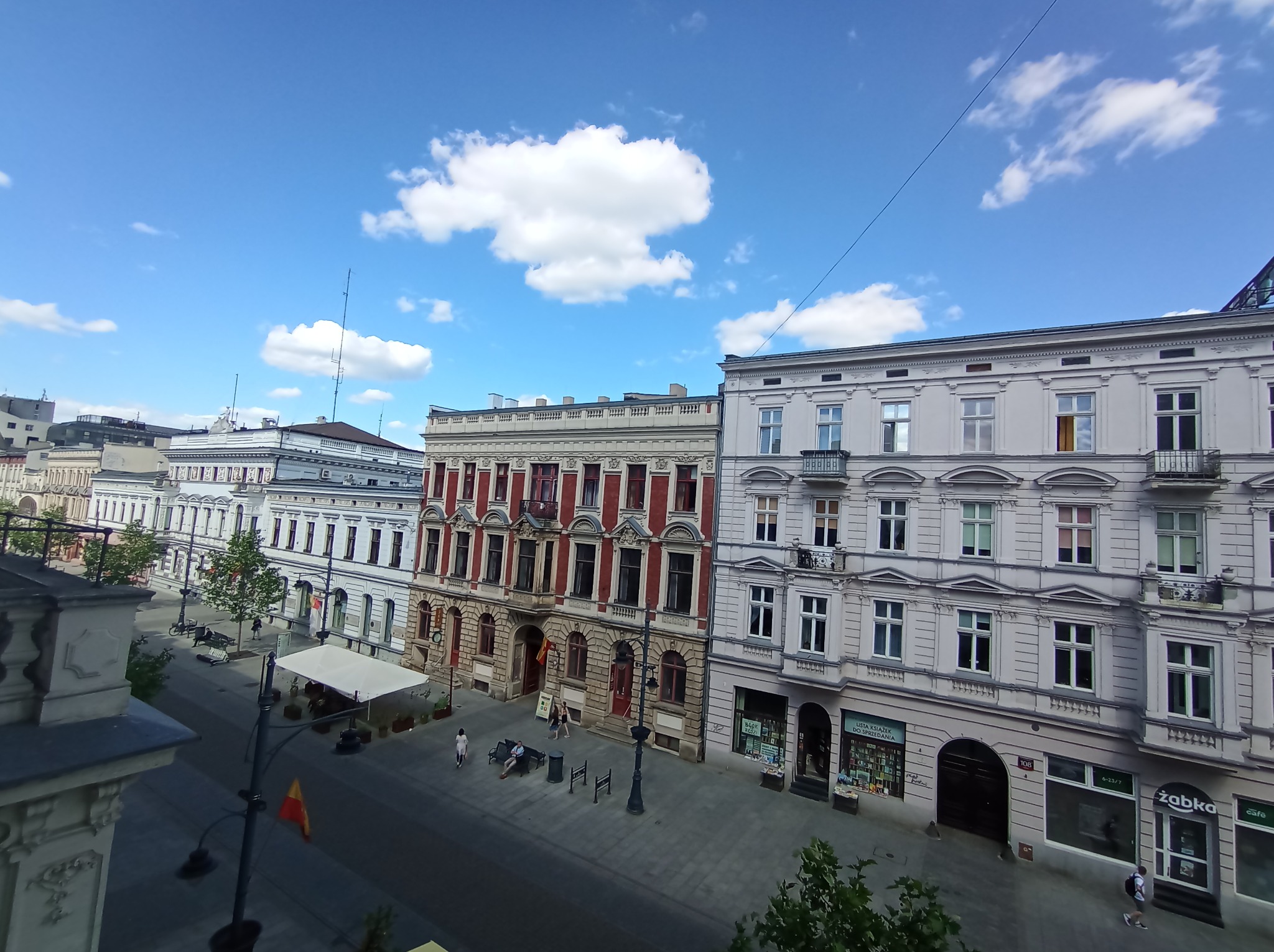 Ulica miejska z budynkami o zróżnicowanej architekturze, widok z góry w słoneczny dzień z błękitnym niebem i białymi chmurami. Na pierwszym planie fragment balkonu.