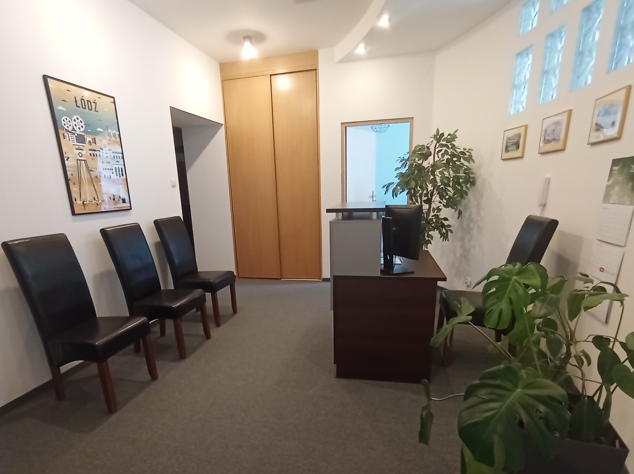 Poczekalnia w kancelarii z ciemnymi krzesłami, recepcją, roślinami doniczkowymi i plakatem 'Łódź' na ścianie.