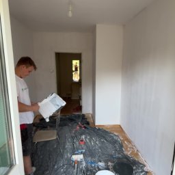 MYSZ-BUD - Mężczyzna z paletą malarską w dłoni, w trakcie malowania ścian na biało. Podłoga zabezpieczona folią, widoczne farby i wałki malarskie. W tle otwarte drzwi.