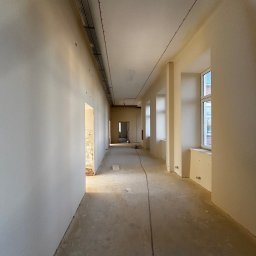 MYSZ-BUD - Długi korytarz w trakcie prac wykończeniowych. Ściany po szpachlowaniu, przygotowane do malowania. Widoczne okna i instalacje pod sufitem. Surowa podłoga.