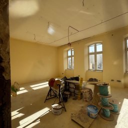 MYSZ-BUD - Wnętrze w trakcie prac wykończeniowych: ściany po szpachlowaniu i malowaniu na kremowo, widoczne narzędzia, kable i materiały budowlane. Jasne światło wpada przez okna.