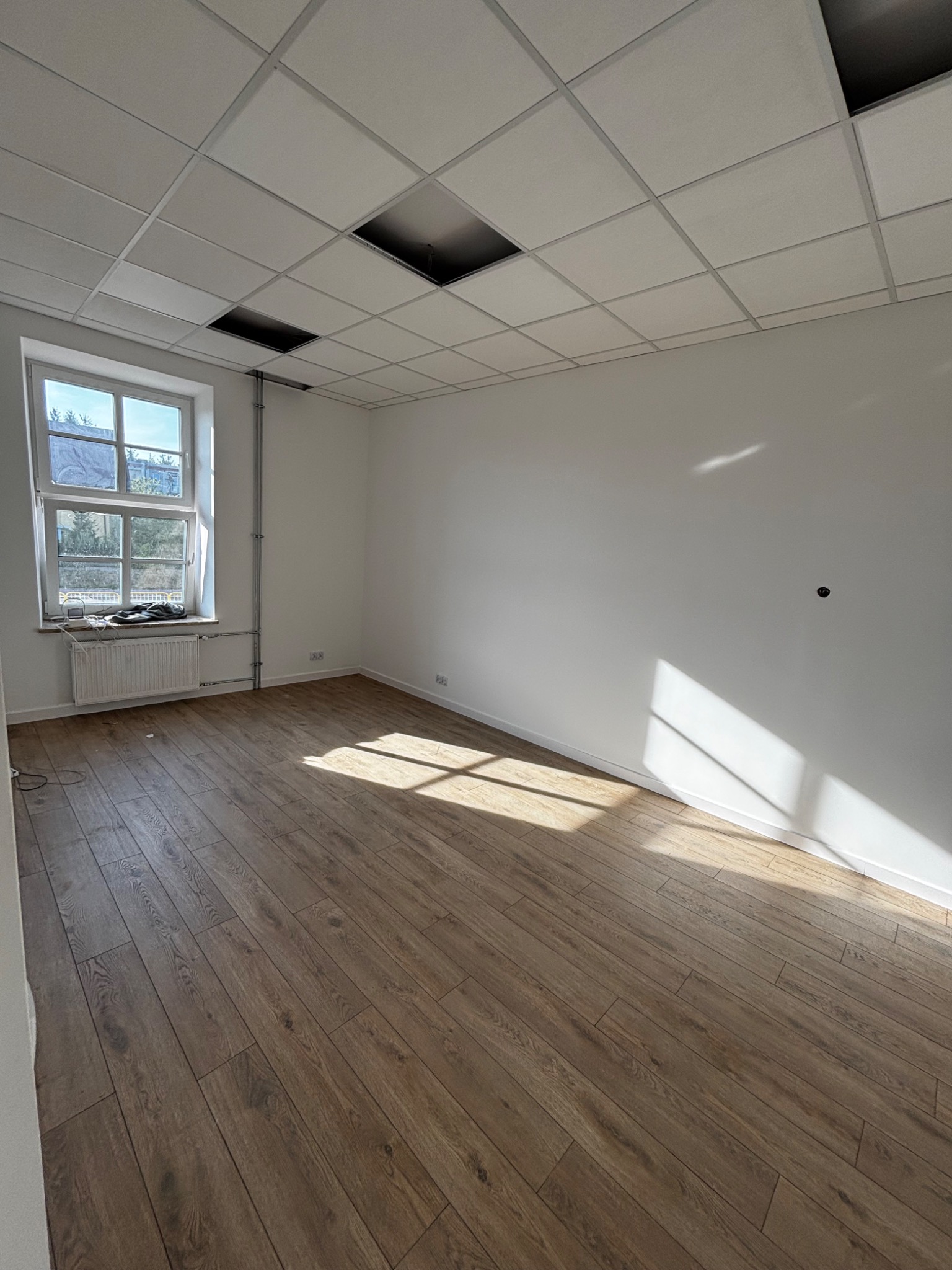 Jasne, puste wnętrze biurowe z drewnianą podłogą, białymi ścianami i kasetonowym sufitem. Okno z radiatorem po lewej stronie. Promienie słoneczne wpadają do pomieszczenia.
