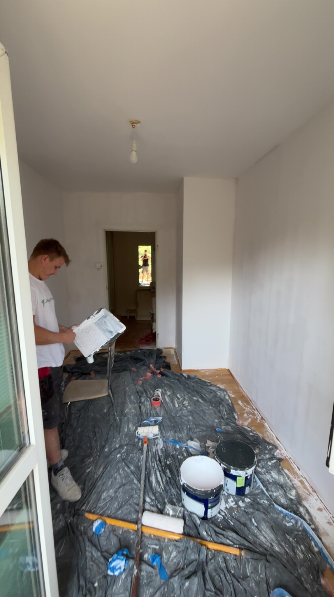 Mężczyzna z paletą malarską w dłoni, w trakcie malowania ścian na biało. Podłoga zabezpieczona folią, widoczne farby i wałki malarskie. W tle otwarte drzwi.