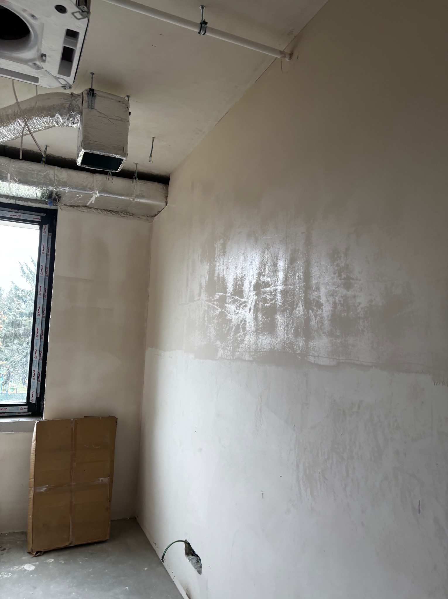 Ściana po szpachlowaniu, widoczne ślady po narzędziach, przygotowana do malowania. W tle okno i karton. Elementy instalacji pod sufitem.