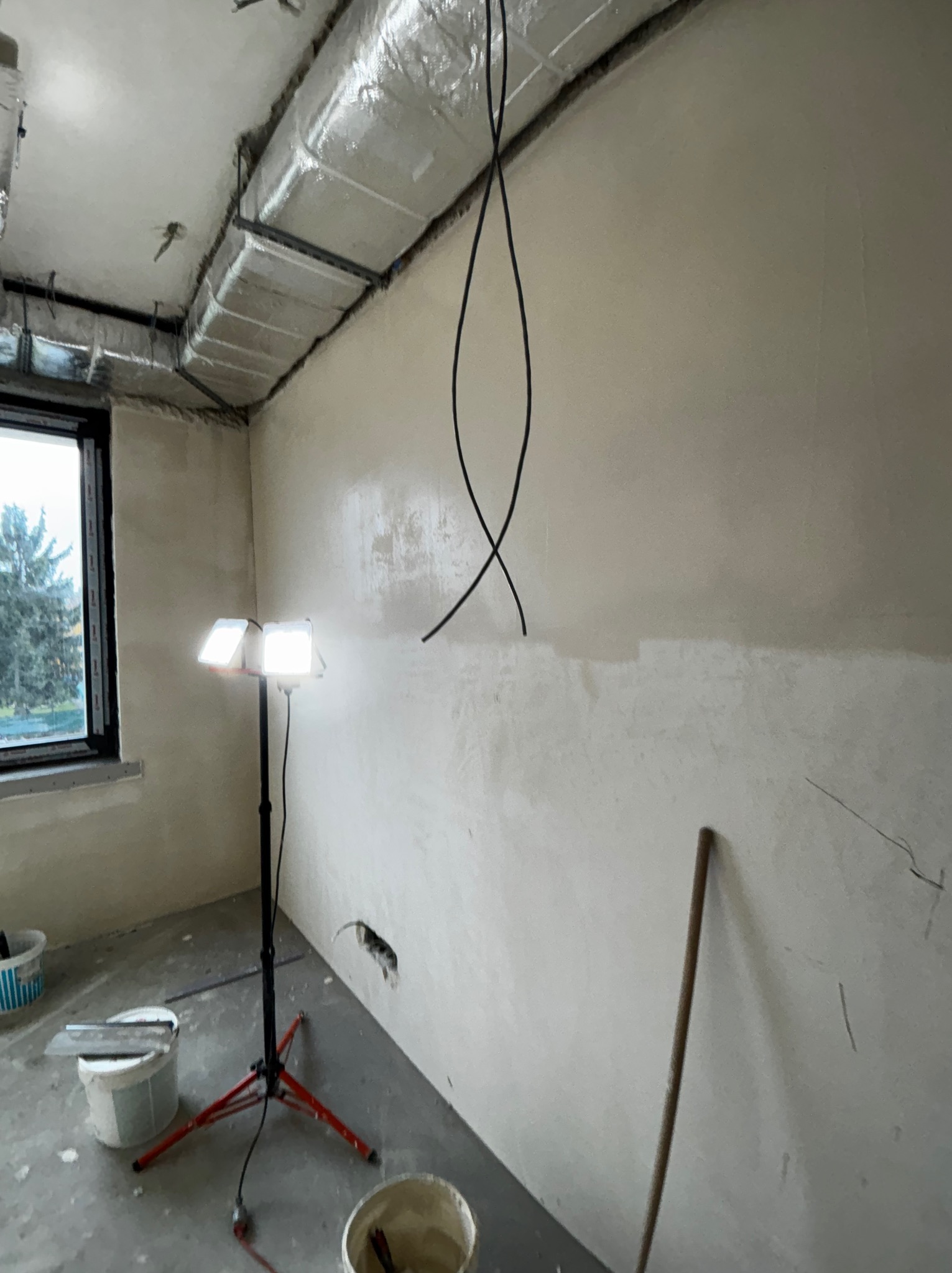 Wnętrze w trakcie szpachlowania: ściany pokryte gładzią, widoczne narzędzia, lampa oświetleniowa, instalacja wentylacyjna pod sufitem. Okno z widokiem na zewnątrz.