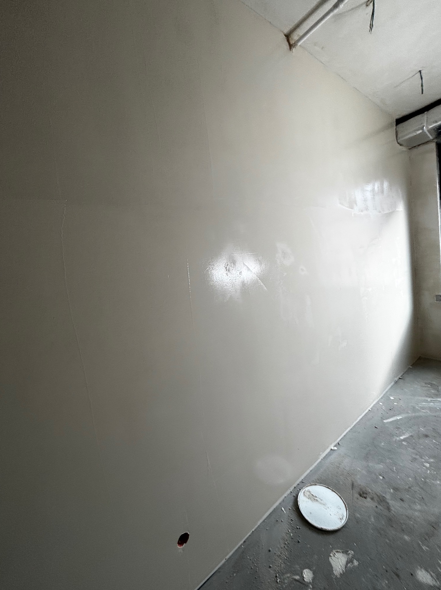 Świeżo wygładzona, jasna ściana w surowym wnętrzu, z widocznymi śladami po szpachlowaniu i niedoskonałościami na podłodze. Białe wieczko po farbie leży przy ścianie.