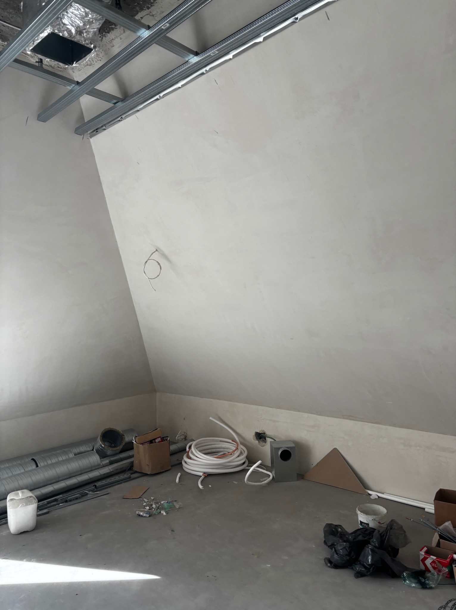 Poddasze w trakcie wykończenia: gładzie na skośnych ścianach, metalowa konstrukcja sufitu, rury wentylacyjne i kable na podłodze. Widoczne niedoskonałości i surowy stan.