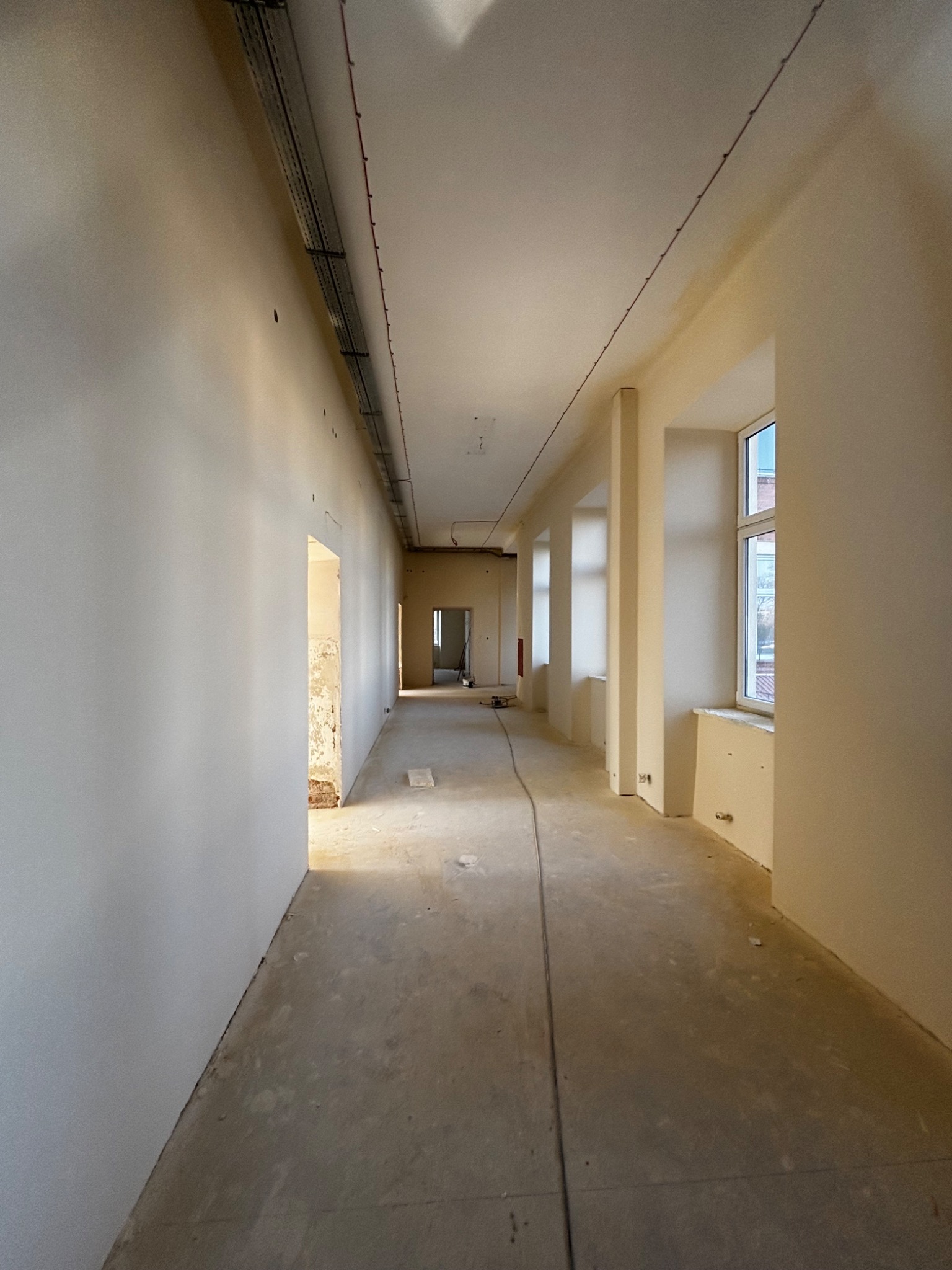 Długi korytarz w trakcie prac wykończeniowych. Ściany świeżo pomalowane na jasny kolor, widoczne instalacje pod sufitem i surowa podłoga. W tle otwarte drzwi.