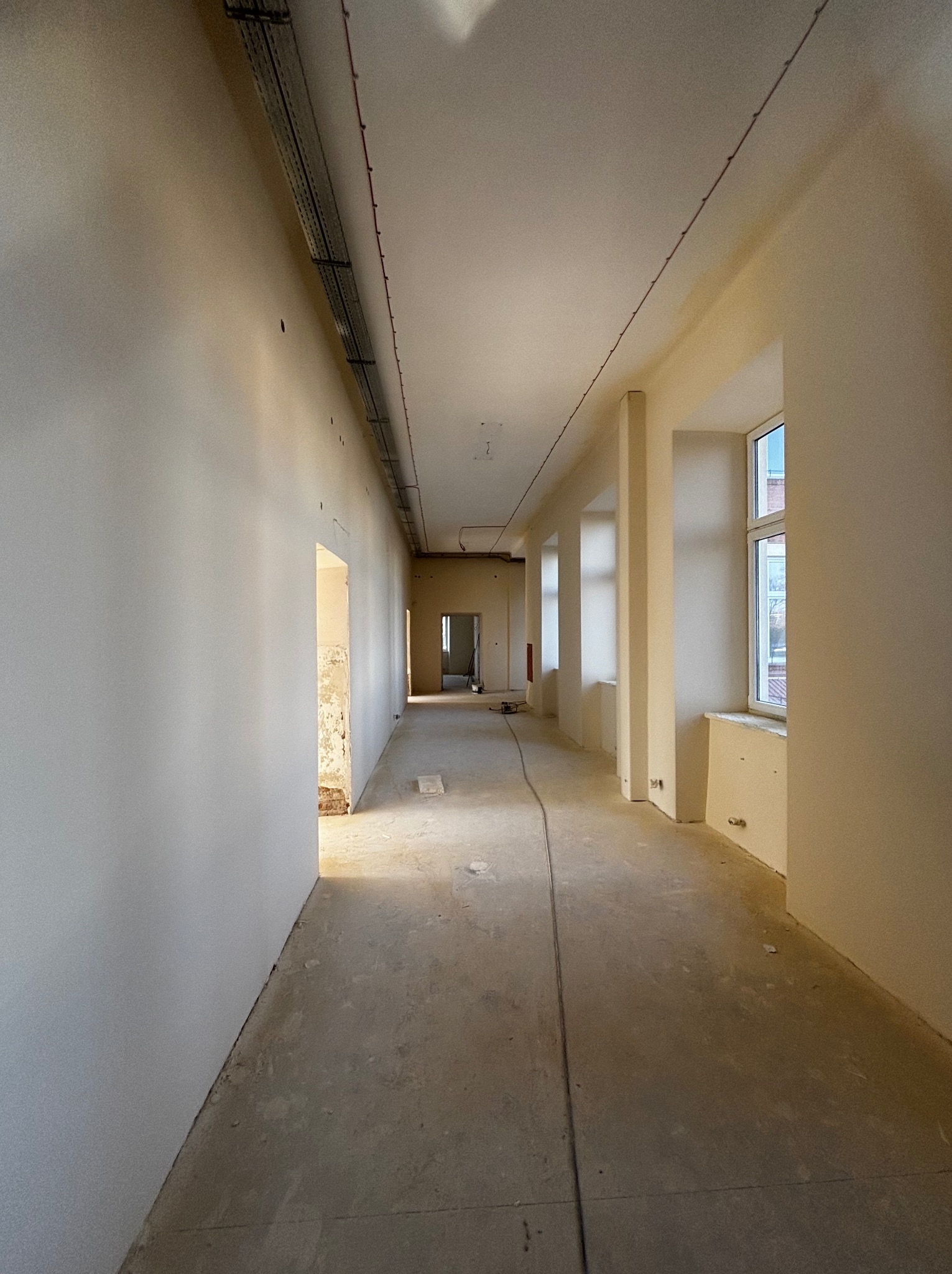 Długi korytarz w trakcie prac wykończeniowych. Ściany po szpachlowaniu, przygotowane do malowania. Widoczne okna i instalacje pod sufitem. Surowa podłoga.