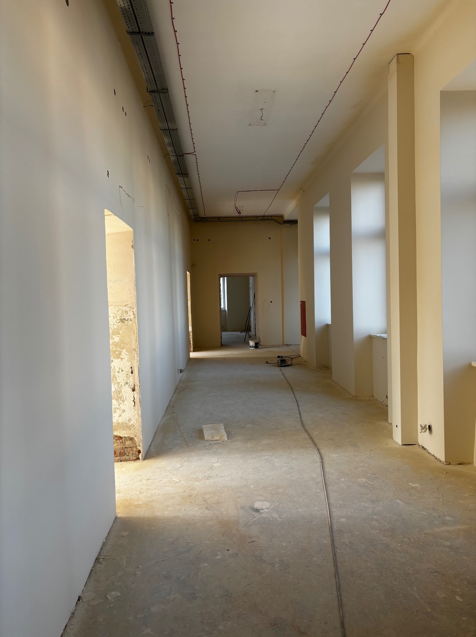 Korytarz w trakcie prac wykończeniowych: świeżo pomalowane ściany na biało, widoczne instalacje pod sufitem, surowa podłoga. Otwory drzwiowe bez ościeżnic.