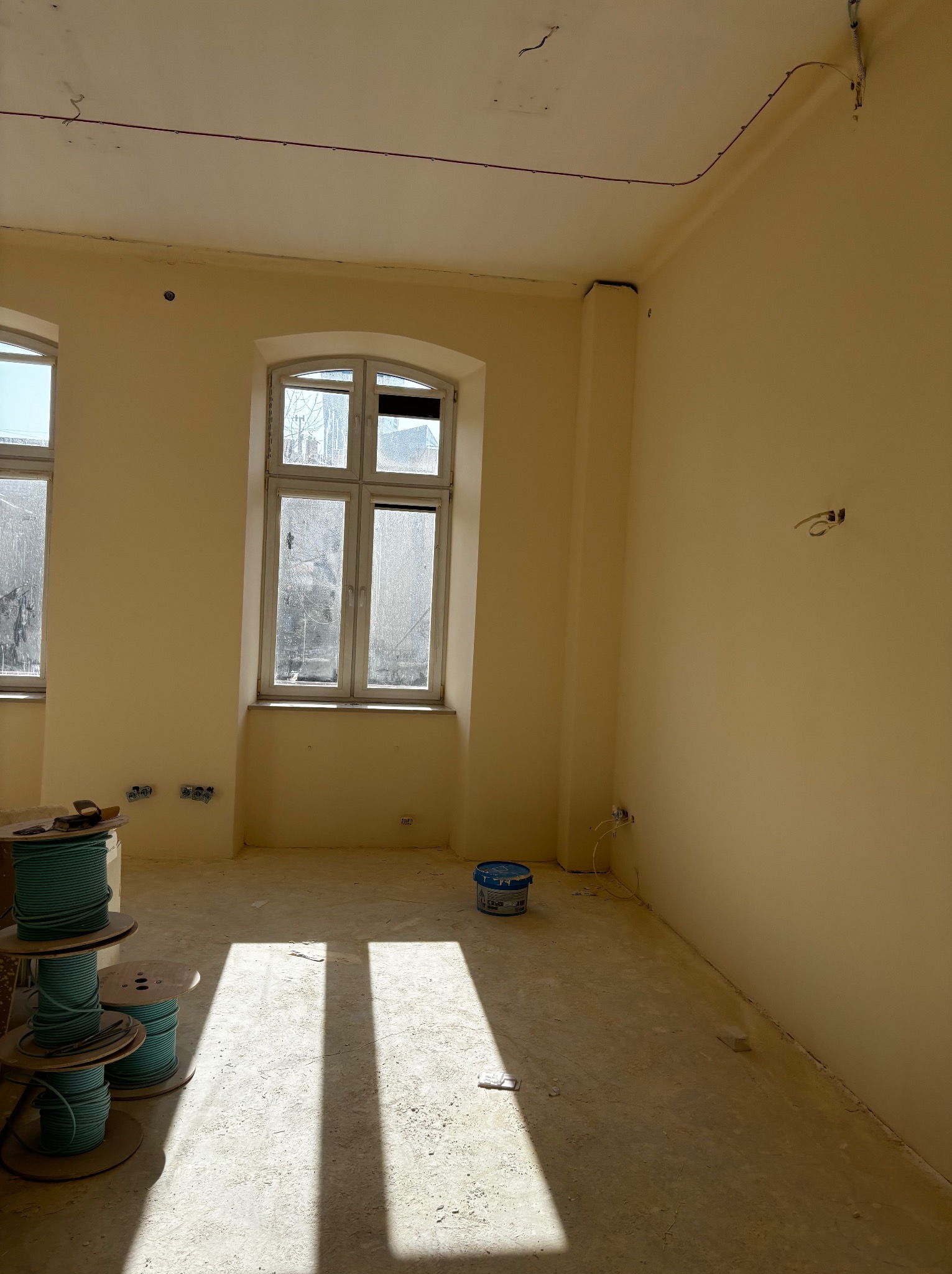 Wnętrze w trakcie remontu: pomalowane ściany na kremowo, widoczne kable elektryczne, puszki, szpule z kablami i wiadro z farbą. Wpadające promienie słoneczne.