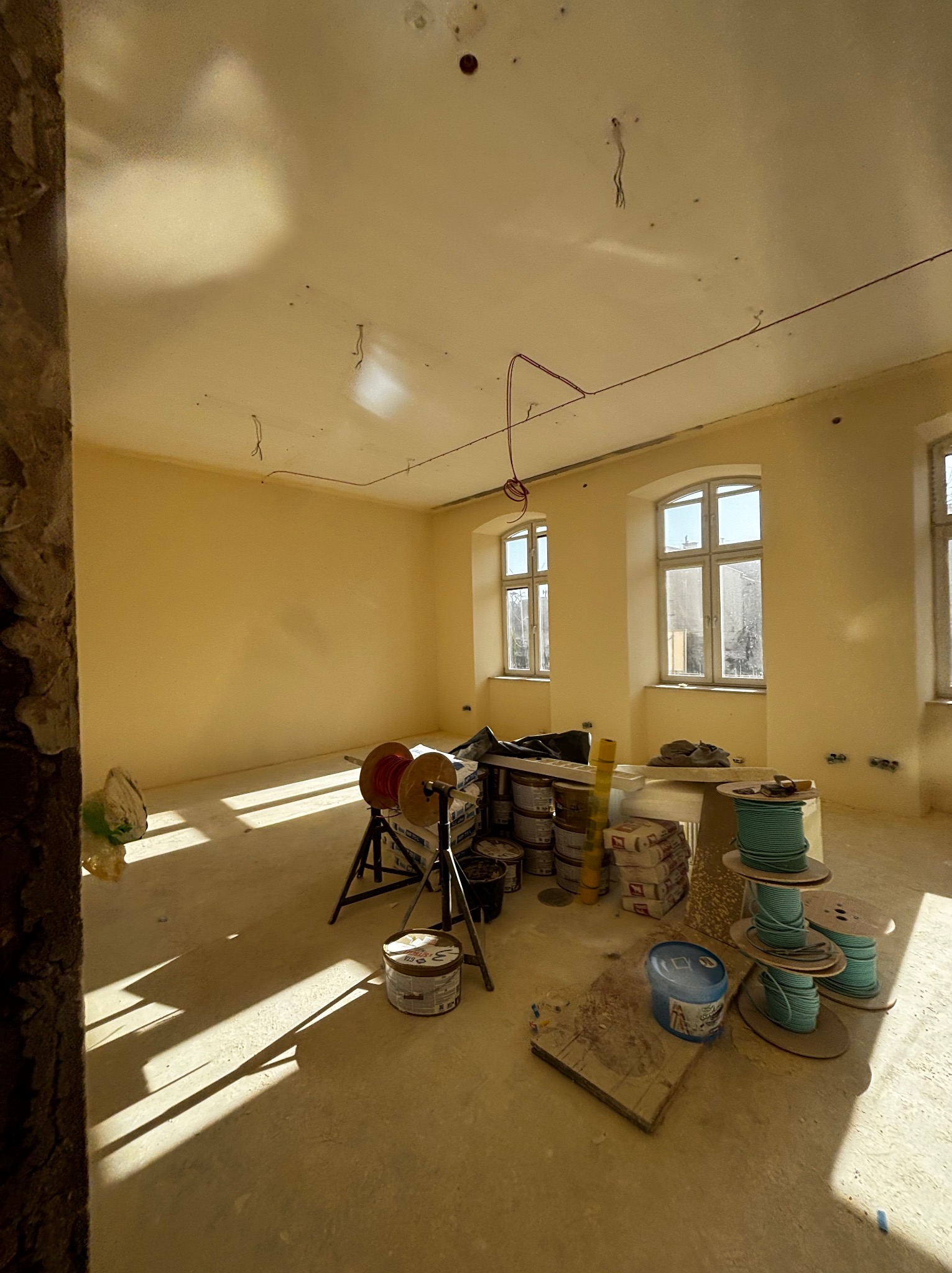 Wnętrze w trakcie prac wykończeniowych: ściany po szpachlowaniu i malowaniu na kremowo, widoczne narzędzia, kable i materiały budowlane. Jasne światło wpada przez okna.