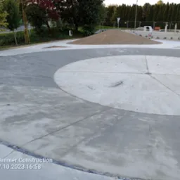 Świeżo wylana, gładka betonowa nawierzchnia placu zabaw z okrągłym wzorem i fragmentem placu ćwiczeń w tle, widoczny kopiec piasku.