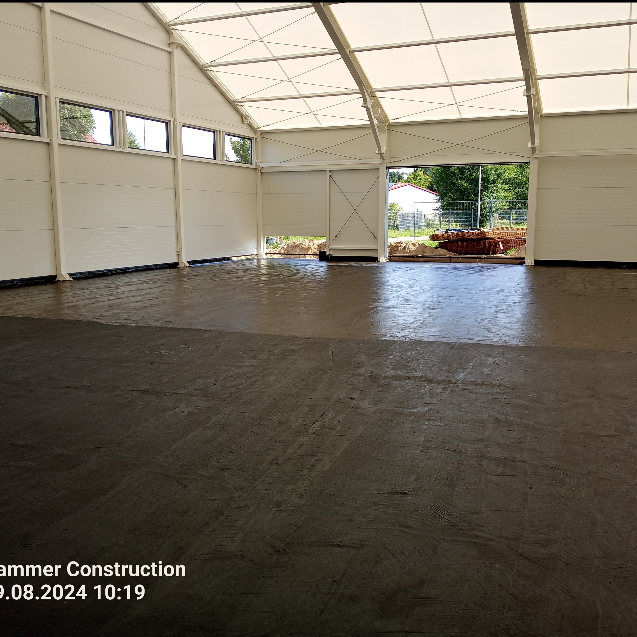 Wnętrze hali produkcyjnej z wylaną, świeżą, ciemną posadzką. Widoczne ślady po rozprowadzaniu masy. Na zewnątrz hali widoczne materiały budowlane i zieleń.