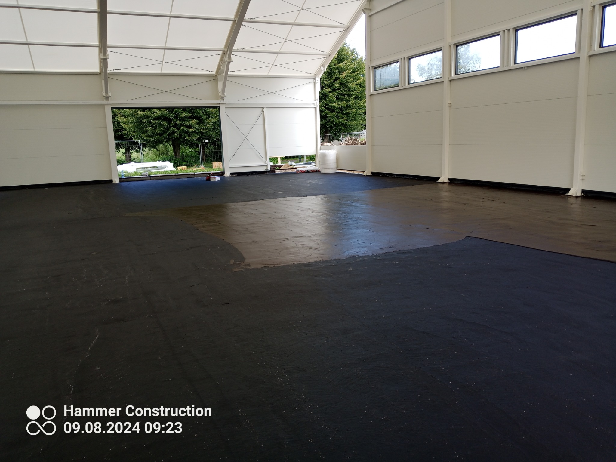 Wnętrze hali w Koszalinie z częściowo wylaną, ciemną posadzką przemysłową, widoczne okna i otwarte wrota hali, w tle zieleń.