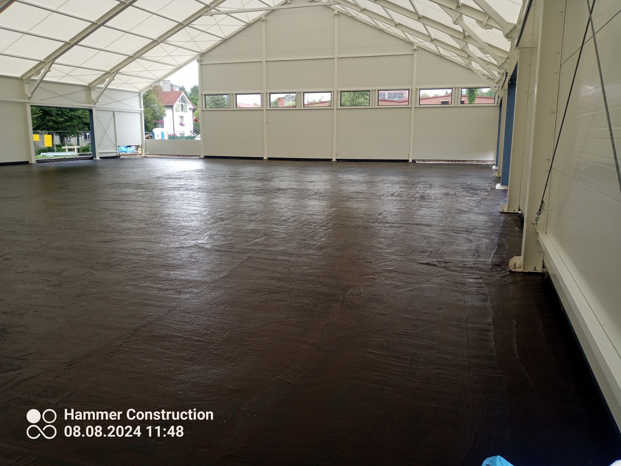 Świeżo wylana, ciemna posadzka w hali magazynowej z widoczną strukturą, białe ściany i konstrukcja dachu, widok na zewnątrz przez okna.